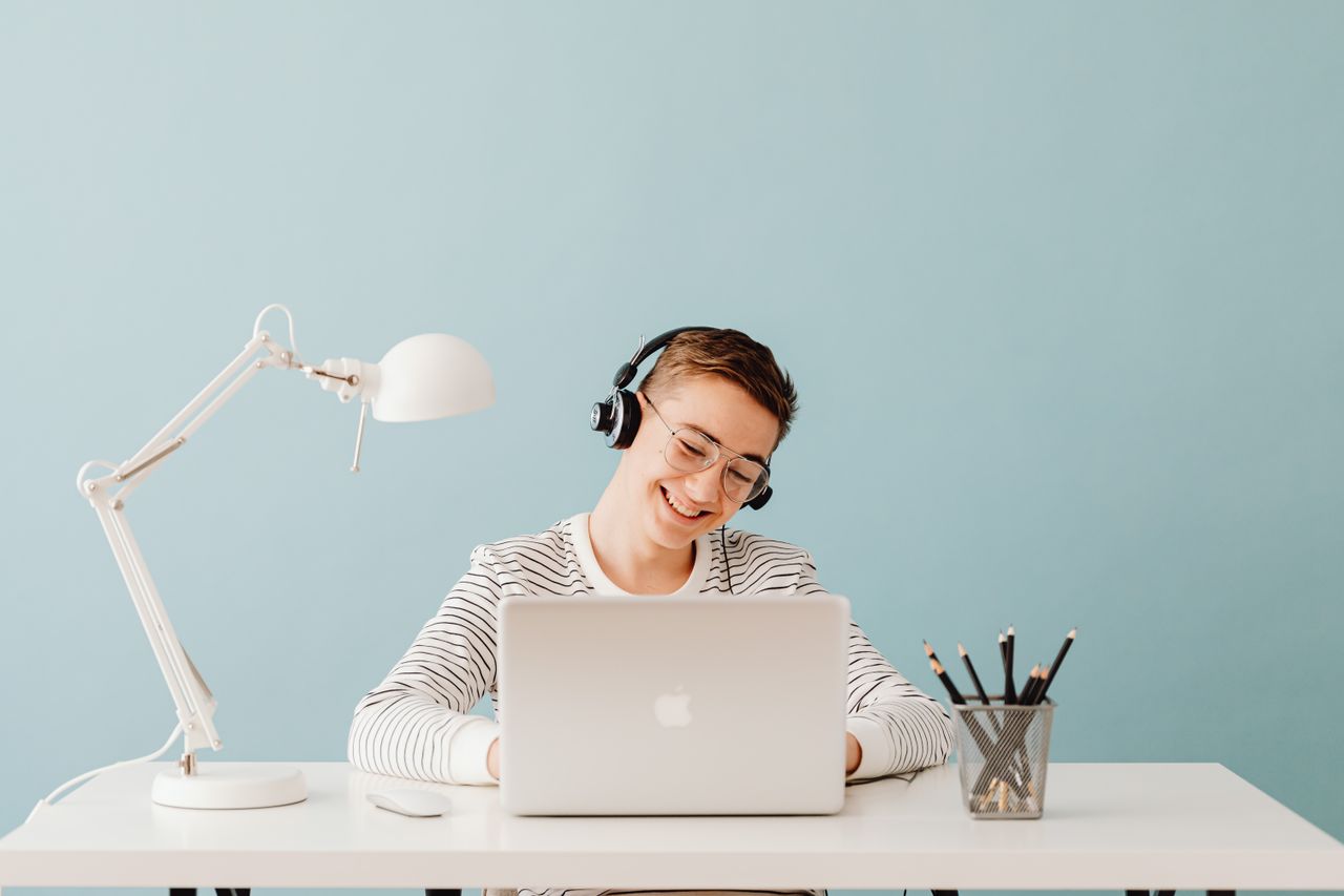 Strona główna A young man smiles while working on a laptop, wearing headphones at a tidy desk with a lamp.